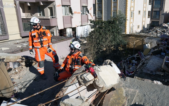 Zwei Männer in orangen Kleidern stehen auf den Trümmern eines eingestürzten Hauses.