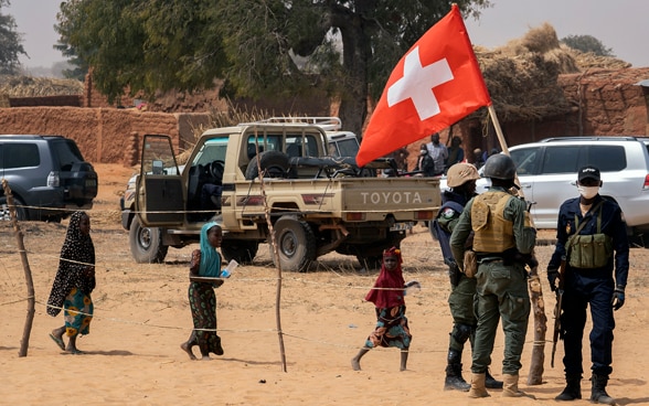  Einige Soldaten stehen neben einem Auto. Eine Person hält eine Schweizer Flagge hoch.