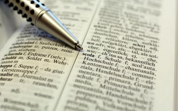 Un dizionario di romancio aperto, con una penna che indica la parola «scola», che significa scuola in romancio.