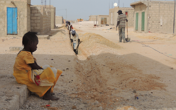 Des habitants de Nouakchott creusent un long sillon rectiligne à la pelle dans une rue sableuse afin d’y poser un tronçon des 50 kilomètres de nouvelles conduites d’eau dans la capitale de la Mauritanie, pendant qu’une petite fille les observe assise sur le bord du trottoir.