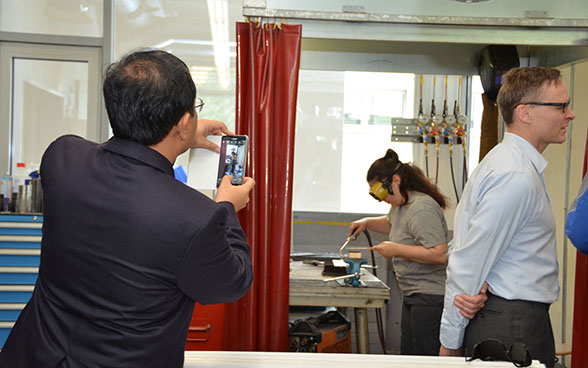 A member of a foreign delegation takes a photo of a young woman welding. 