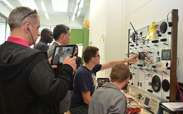 Two car diagnostics technicians work with cables, switches and speakers on an infotainment systems wall. Behind them are the members of a foreign delegation on a visit to the Swiss Technical College in Winterthur.
