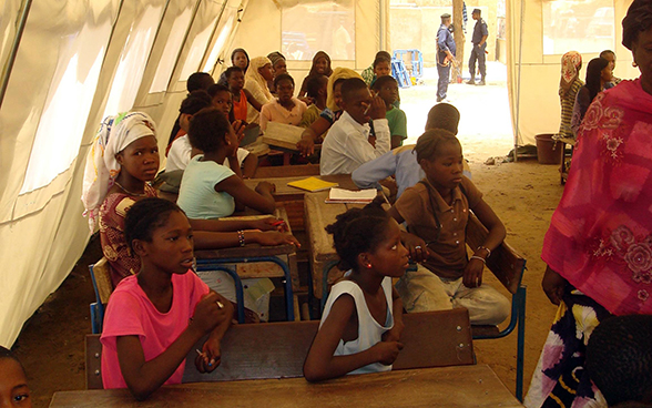 Des enfants sont assis derrière des bancs d’école installés sous une tente.