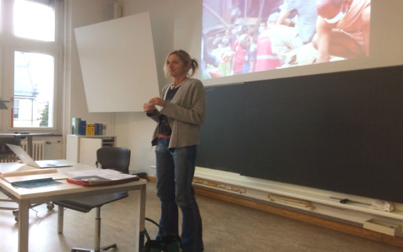 Eine Frau steht in einem Schulzimmer zwischen einem Tisch und einer Wandtafel. Über der Tafel ist ein Bild an die Wand projiziert. 