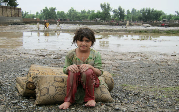 Ein Mädchen in Pakistan sitzt auf einem Kissen im Freien. Hinter dem Mädchen befindet sich eine Wasserlache. 