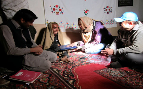 Eine Frau und zwei Männer sitzen in einem Zelt, die Frau überreicht dem einen Mann einen Laptop.