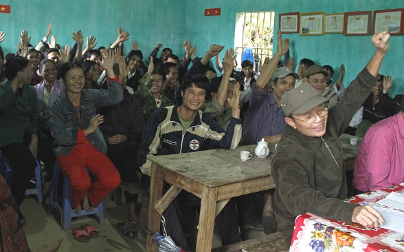 Dorfbewohnerinnen und Dorfbewohner in einem Raum stimmen durch Handaufheben ab.