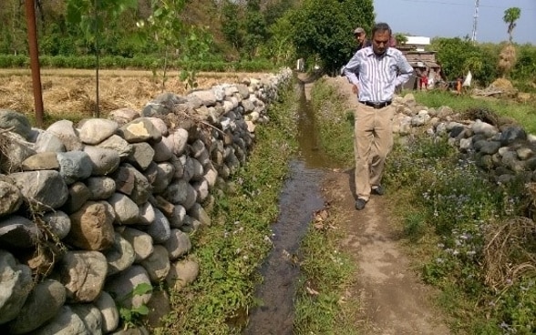 Zwei Männer schreiten einen Bewässerungskanal, der entlang einer Steinmauer fliesst, ab, um dessen Zustand zu evaluieren.