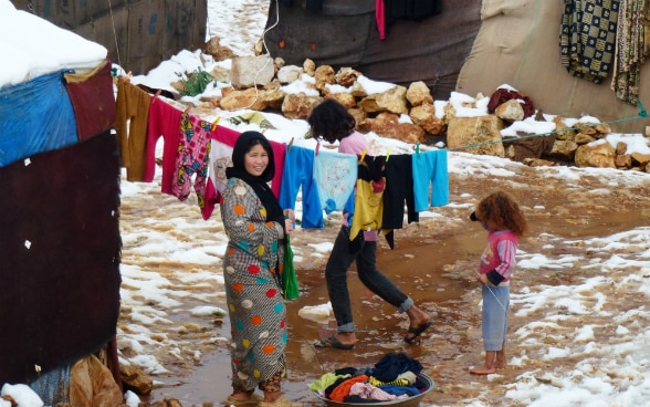 Eine Frau in einem Flüchtlingslager beim Aufhängen der Wäsche zwischen zwei Zelten. Daneben stehen zwei Kinder. Am Boden liegt Schnee.
