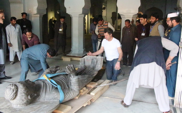 La statua di Buddha viene disposta su pallet in legno per essere portata fuori dalla sala espositiva. Molte persone assistono alla scena.
