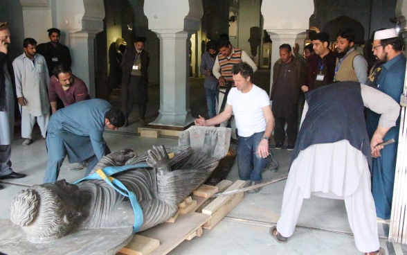 Die Buddha-Skulptur wird auf hölzerne Paletten gelegt, damit sie den Ausstellungssaal verlassen kann. Zahlreiche Personen stehen darum herum und beobachten die Szene.