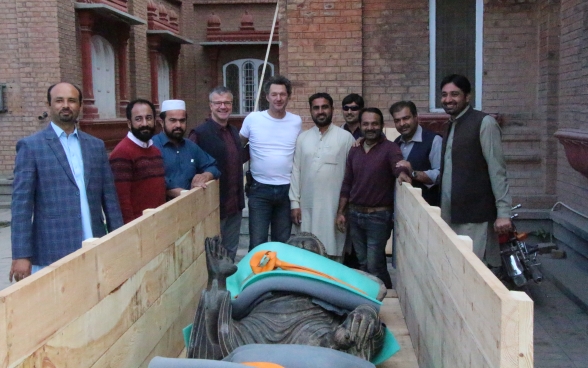 La statua di Buddha è posizionata in un contenitore di legno aperto ed è fissata con varie cinghie. Attorno alla cassa posano i due rappresentanti svizzeri e il team pakistano.