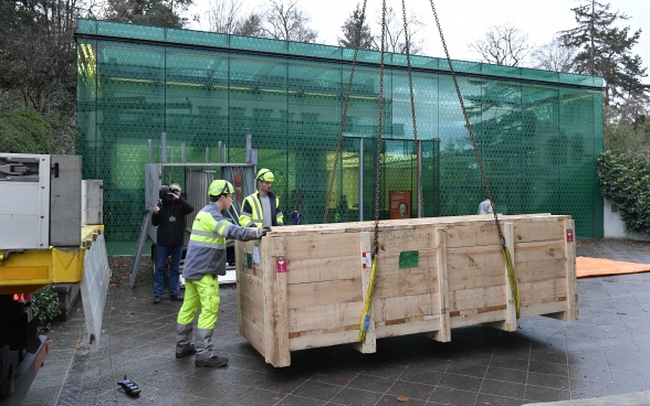 Die Transportkiste hängt an Ketten an einem Kran und wird vor dem gläsernen Bau des Museums Rietberg abgeladen.