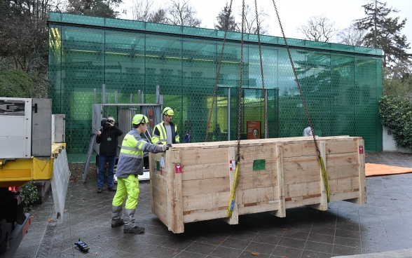 Il contenitore per il trasporto, fissato a una gru mediante catene, viene posizionato davanti all’edificio di vetro del Museo Rietberg. 