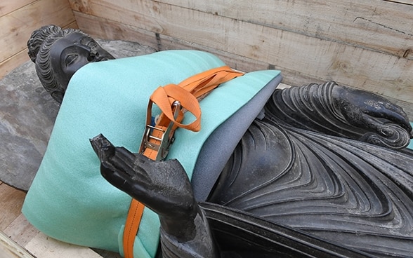 Una statua in pietra raffigurante Buddha è fissata con una cinghia all’interno di un contenitore di legno adibito al trasporto.
