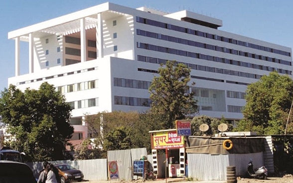 Das Krankenhaus in Pune ist ein grosses weisses Gebäude umringt von Bäumen. Im Vordergrund stehen Holzbaracken.