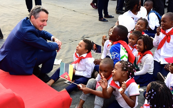 Il consigliere federale Cassis è seduto su un sofà rosso e parla con bambini di una scuola africana