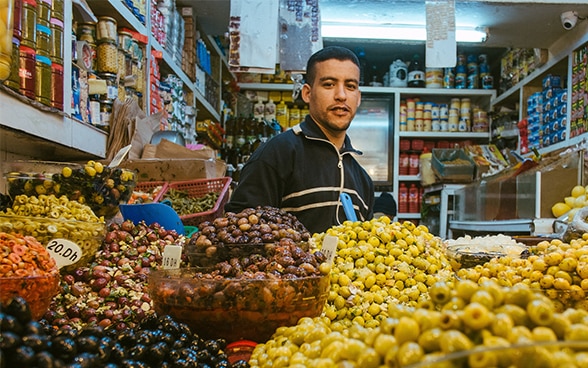 In un piccolo negozio un uomo è in piedi davanti a delle grandi ceste colme di olive.