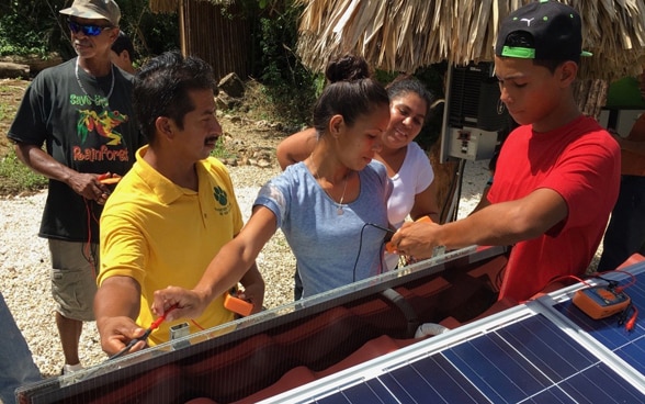 Men and women from the local community stand near a roof with solar panels and are taught the basics of how to use them.