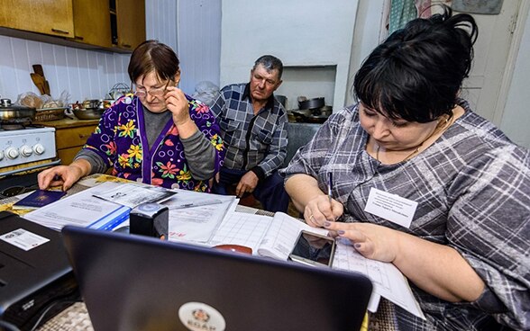 Dos mujeres y un hombre sentados en una cocina, leyendo documentos frente al ordenador. 