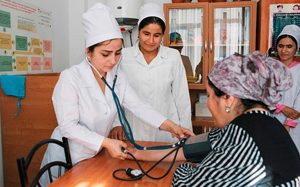 Two Tajik doctors measure the blood pressure of a patient sitting on a chair.