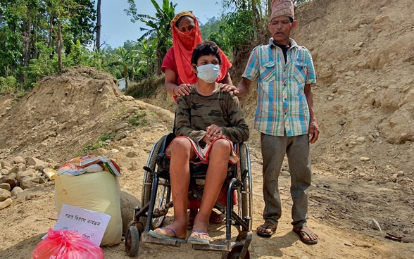 Eine Frau und ein Mann stossen einen jüngeren Mann in einem Rollstuhl durchs Gelände in Nepal.