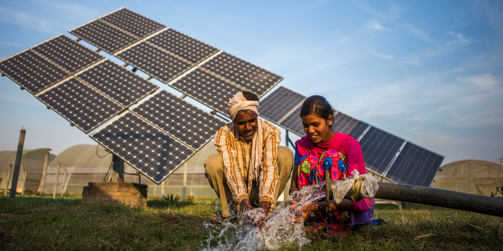 A man and woman in the Global South sit at a water pipe in front of an array of solar panels.