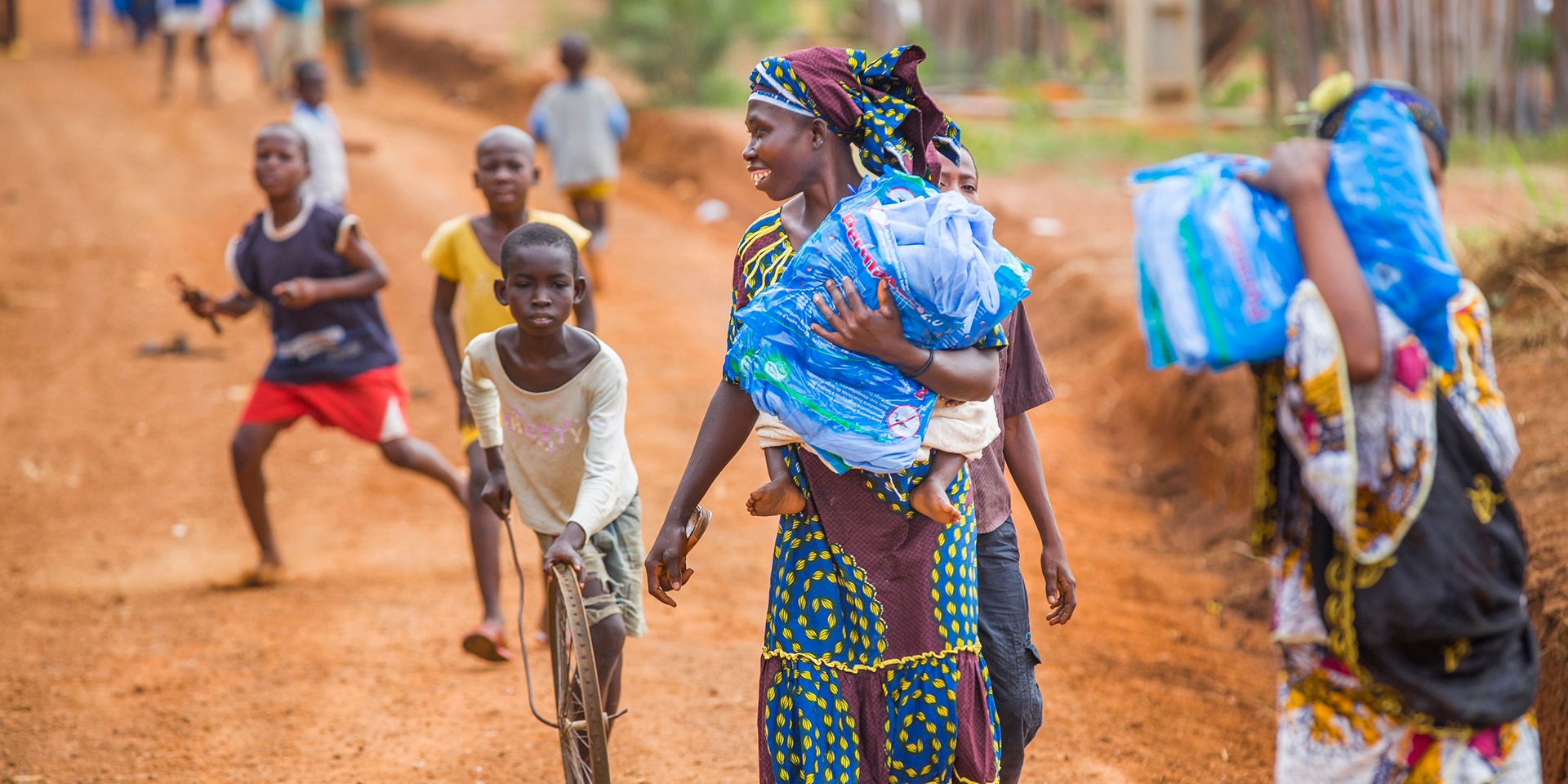 A woman in the foreground is walking with mosquito nets in her hands. Behind her are children.