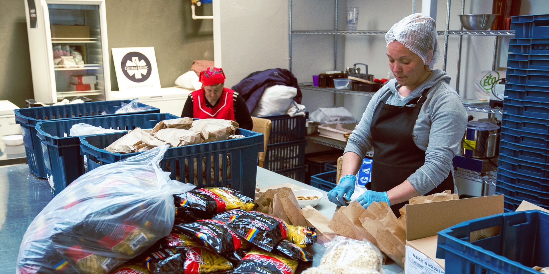 Two women are packing food. Packages of pasta lie on a table in the foreground.