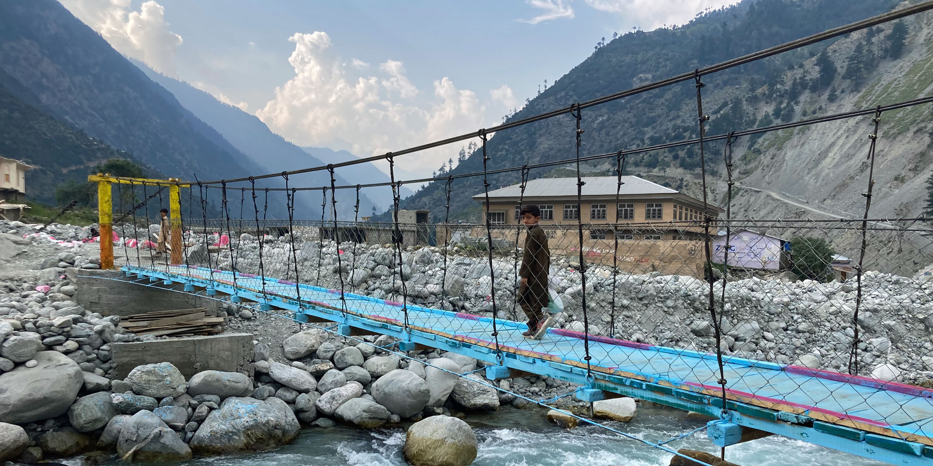 Un garçon traverse un pont de corde près de Mankiala, au Pakistan. Une étendue de grosses pierres borde un ruisseau.