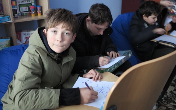 A boy sits in a room and writes on a piece of paper on a chair in front of him. Around him, other children are learning in a similar setup. 