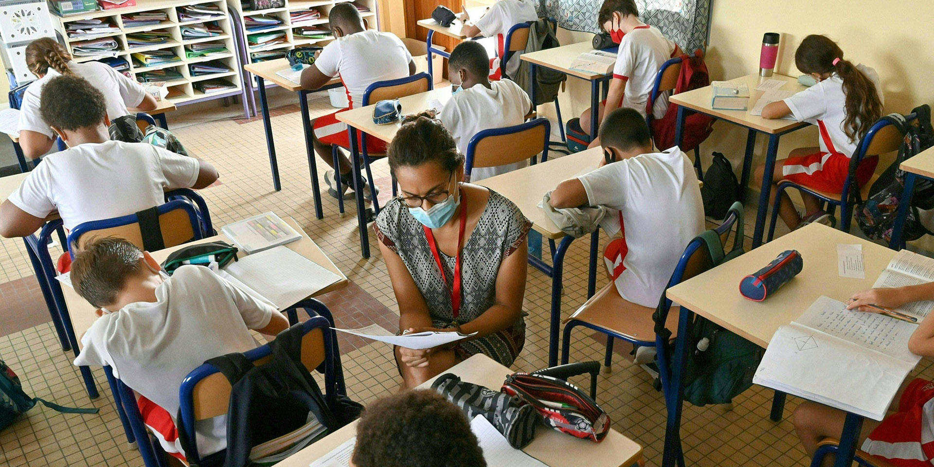 Dei bambini sono seduti tra i banchi di scuola. La maestra aiuta a svolgere i compiti.