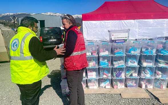 Eine Frau des Schweizerischen Korps für humanitäre Hilfe in einer roten Weste sowie ein Mann mit einer gelben Weste stehen vor Kisten mit Hygienepaketen.