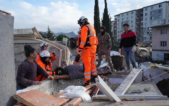 Four men lifting a stretcher with a person on it out of the ruins of a collapsed house.