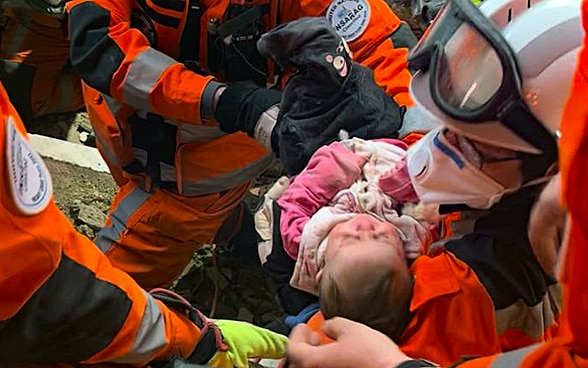 TEXT Ein Baby liegt in den Armen einer Frau mit Helm und orangen Kleidern, um sie herum zwei weitere Mitglieder der Rettungskette.