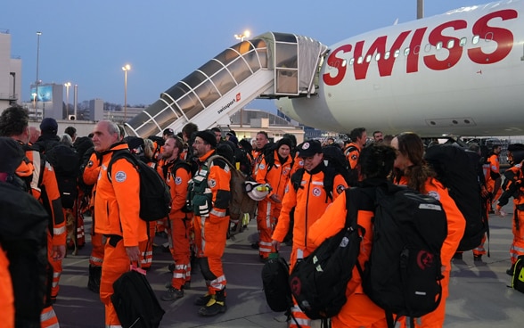 Numerous members of the rescue chain in orange clothing stand on an airfield. In the background you can see an aircraft with a staircase docked to it.