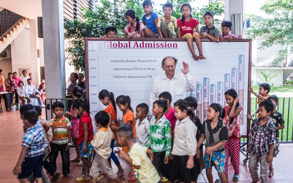 Beat Richner photographié en 2013 au Cambodge, au milieu des enfants. Derrière lui, un tableau avec un graphique présentant le nombre de petits patients soignés chaque année dans les hôpitaux.