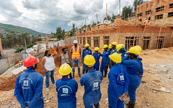 Mujer conversando con un grupo de personas vestidas con overoles azules, todas de pie formando un círculo. En el fondo, trabajadores construyendo una casa de ladrillo.
