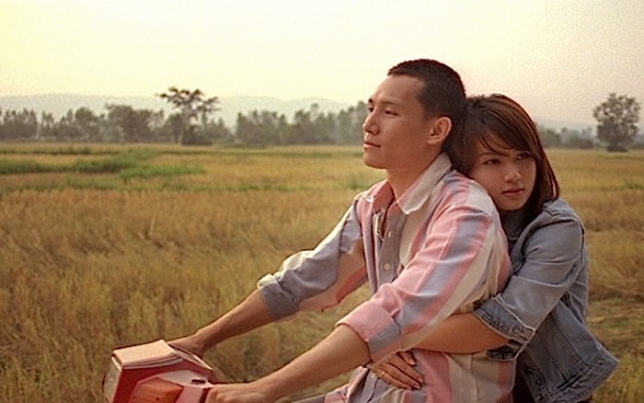 A couple riding a scooter, with the man in front and the woman behind him with her arms wrapped tightly around him.