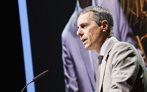 Federal Councillor Ignazio Cassis standing behind a lectern on stage. The screen on which the speech is being transmitted can be seen in the background.