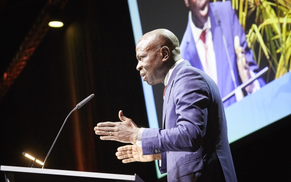 Gilbert F. Houngbo est debout sur la scène devant le pupitre. En arrière-plan on voit un grand écran sur lequel est projeté le discours.