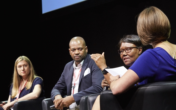 Andrea Heinzer, Sithole Mbanga, Malijeng Ngqaleni and Melanie Pfändler sitting on stage. Malijeng Ngqaleni speaking into a microphone.