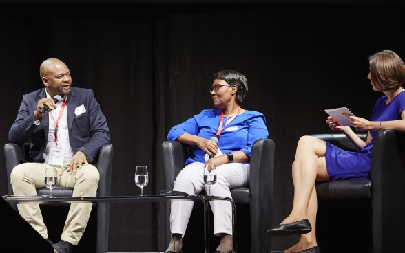 Malijeng Ngqaleni e Melanie Pfändler siedono sul palco e ascoltano Sithole Mbanga, seduto di fianco a loro, mentre parla al microfono.