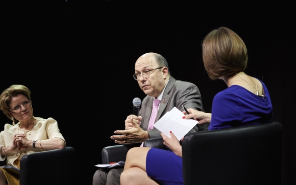 Marie-Gabrielle Ineichen-Fleisch and Melanie Pfändler sitting on stage listening attentively to Thomas Gass, sitting in the middle and speaking into a microphone.