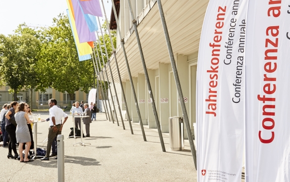 La Festhalle de Bernexpo depuis dehors. Des drapeaux colorés sont suspendus devant les portes et sur des bannières on peut lire en plusieurs langues nationales et en anglais : « Conférence annuelle ». 