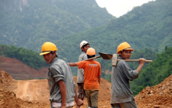 Four young men wearing helmets, carrying a shovel and a sledgehammer, surrounded by earthworks with a forest rising in the background.