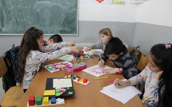 Girls and boys are in their classroom sitting around a table in front of the blackboard. They each make a coloruful drawing. 