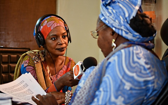 An African woman holds a microphone as she interviews another woman in a radio studio.