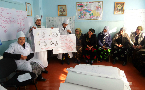 Tre operatrici in camice bianco trasmettono informazioni sull’allattamento a un gruppo di donne tagiche con l’aiuto di cartelloni.