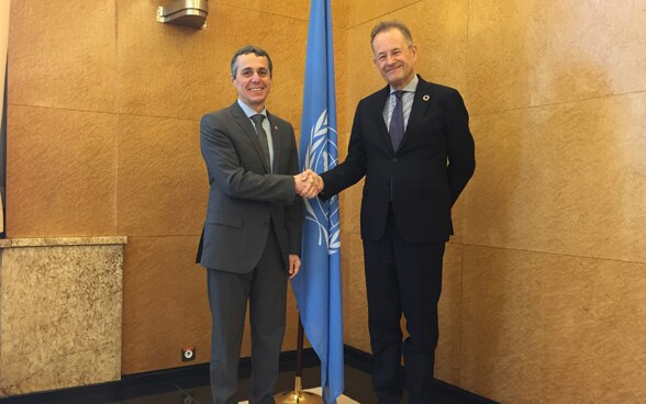 Le conseiller fédéral Ignazio Cassis et Michael Moller, directeur des Nations Unies à Genève, se serrent la main devant un drapeau des Nations Unies.
